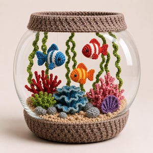 May include: A glass fishbowl featuring a crocheted brown rim and base, filled with three small crocheted fish in blue, red, and orange. The interior includes crocheted green seaweed, red and pink coral, a blue anemone, and a purple shell.