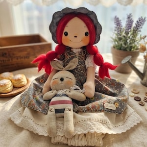 Puede incluir: Una muñeca de tela con cabello de lana rojo brillante en trenzas y un gorro gris oscuro, que viste un vestido con estampado floral sobre una falda color crema con ribete de encaje. La muñeca sostiene un pequeño conejo de peluche beige.