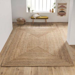 May include: A rectangular area rug crafted from natural tan jute fibers, featuring a woven geometric border pattern that creates a central diamond shape. The rug is laid flat on a light-colored wooden floor in a bright, open room setting.