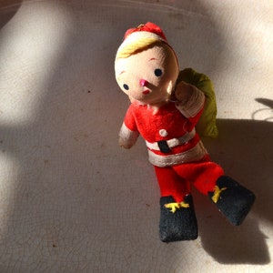 May include: A vintage Christmas ornament depicting a Santa Claus doll wearing a red and white suit with a black belt and a yellow bag. The doll has a painted face with rosy cheeks and black eyes.