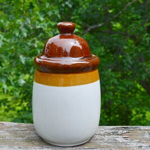 May include: A white ceramic jar with a brown and orange lid. The jar has a smooth, rounded shape and a glossy finish.