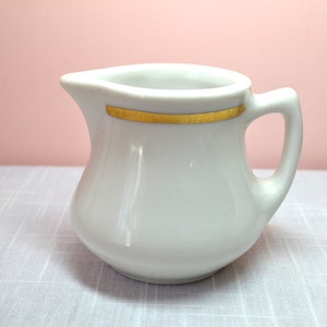 May include: White ceramic creamer with a gold band around the top. The creamer has a rounded body and a small handle.