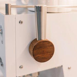 Puede incluir: Una máquina de café espresso blanca con una palanca vertical prominente, un mango de plata pulida y un detalle circular de madera marrón oscura. La máquina incluye una serie de pequeños interruptores de palanca plateados y un riel metálico.