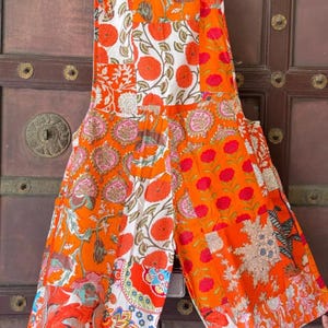 Könnte beinhalten: Eine Latzhose im Patchwork-Stil mit einem lebendigen Blumenmuster in Orange, Weiß und Pink. Das Kleidungsstück besteht aus mehreren Stoffbahnen mit unterschiedlichen botanischen Mustern, darunter große kreisförmige Blüten, Ranken und komplexe Paisley-Motive.