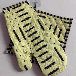 Puede incluir: Un guante de cocina acolchado y un agarrador cuadrado a juego en tela de color amarillo pálido con un patrón abstracto negro de líneas onduladas y puntos. Ambos artículos tienen un borde de tela a rayas blancas y negras en los extremos.
