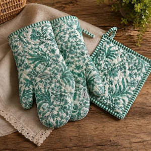 May include: A pair of quilted oven mitts and a matching square pot holder featuring a white base with a detailed green floral and botanical print. Each piece is finished with a green and white striped fabric trim along the edges.