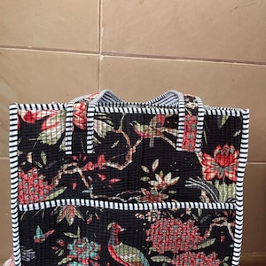 Puede incluir: Un bolso tote acolchado rectangular con fondo negro y un patrón colorido de flores y pájaros. El bolso tiene dos asas y un bolsillo frontal, todo rematado con un borde de tela a rayas blancas y negras a lo largo de los bordes y costuras.