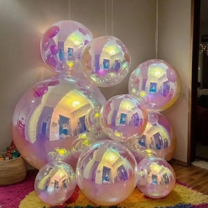 Op de afbeelding: Een verzameling ronde, iriserende ballonnen in verschillende maten, met een reflecterend, halfdoorzichtig oppervlak dat een regenboogachtige glans vertoont. De ballonnen zijn gegroepeerd en vormen een verzameling van bolvormige objecten.