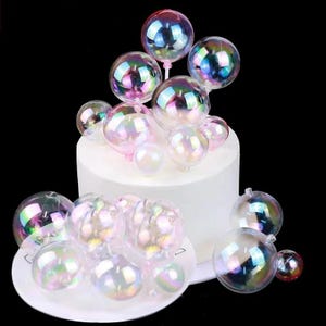 May include: A collection of iridescent, translucent spherical ornaments in varying sizes. Several spheres are attached to thin white stems and arranged atop a cylindrical white cake, while others are clustered on a small white plate nearby.
