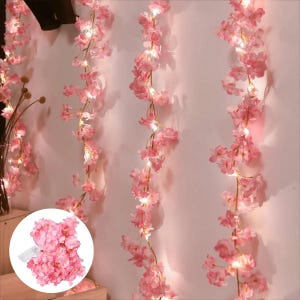Puede incluir: Una guirnalda de flores de cerezo artificiales de color rosa entrelazadas con pequeñas luces LED de color blanco cálido. La guirnalda floral cuelga verticalmente en varios hilos, con pétalos delicados y tallos finos, y una caja de pilas visible.