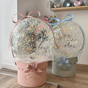 Op de afbeelding: Twee ronde geschenkdozen met doorzichtige ballonstoppers gevuld met pastelkleurige schuimballetjes. De linkerdoos is roze met een bijpassende strik en de naam Emily. De rechterdoos is lichtblauw met een bijpassende strik en de naam Charlie. Gouden Happy Easter tekst.