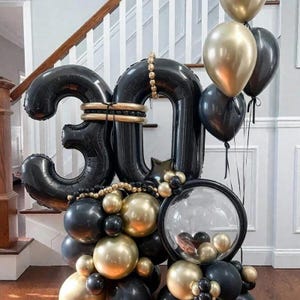 Op de afbeelding: Een ballonarrangement met grote zwarte folieballonnen in de vorm van het getal 30, geaccentueerd met gouden en zwarte latexballonnen van verschillende groottes, inclusief een doorzichtige bubbelballon met kleinere ballonnen erin.
