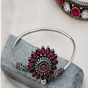 Könnte beinhalten: Ein silberfarbener Metallarmreif mit einem zentralen blumenförmigen Ornament, das mit tiefrosa, blütenblattförmigen Steinen besetzt ist. Das Band weist strukturierte, gewundene Metalldetails in der Nähe des Mittelstücks auf, ein zweiter Armreif ist sichtbar.