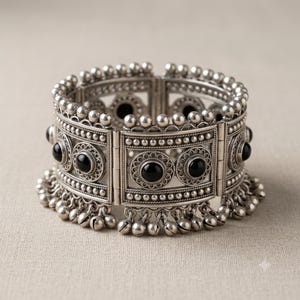 Könnte beinhalten: Ein breites, aufklappbares Armband in Silberoptik mit runden schwarzen Steineinlagen, die von aufwendigen Metallperlen umgeben sind. Die Kanten sind mit kleinen silbernen Kugeln besetzt und der untere Rand ist mit einer Reihe hängender Silberglöckchen verziert.