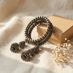 Könnte beinhalten: Ein Armreif in oxidierter Silberoptik mit einem Mittelband aus filigranem Drahtgeflecht und Reihen kleiner Metallperlen. Drei glockenförmige Jhumka-Anhänger mit detaillierten Mustern hängen an der Unterseite des runden Armreifs.