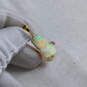 Op de afbeelding: Een hanger van ruwe, ongeslepen opaal met een goudkleurig metalen oogje. De edelsteen heeft een doorschijnende, melkwitte basis met iriserende flitsen van groen, oranje en geel licht over het onregelmatige, getextureerde en organische oppervlak.