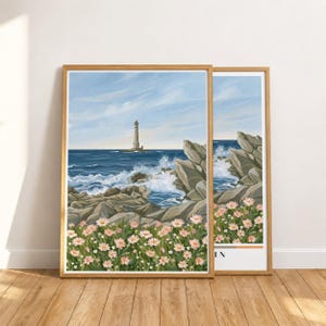 Op de afbeelding: Een ingelijste kunstdruk met een kusttafereel met een vuurtoren in de verte boven een diepblauwe oceaan. De voorgrond toont grillige grijze rotsen en een veld met lichtroze bloemen onder een lichtblauwe lucht met ijle witte wolken.