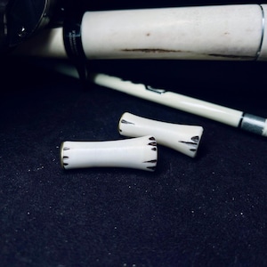 May include: Two small, off-white, cylindrical components with flared ends and dark, etched markings along the edges. These pieces are positioned in front of a fishing rod that features a matching off-white handle and a dark reel assembly.