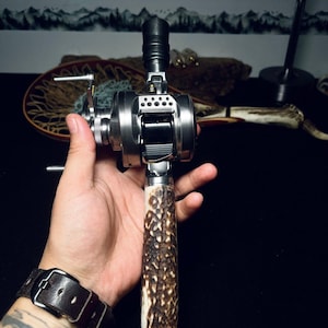 May include: A silver baitcasting fishing reel featuring a textured brown antler handle grip. The reel has a metallic finish with a black spool release lever and a side-mounted crank handle. The design incorporates a perforated metal plate near the top.