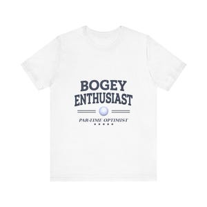 May include: A white short-sleeve t-shirt featuring dark blue text that reads BOGEY ENTHUSIAST above a small white golf ball graphic, with the text PAR-TIME OPTIMIST and five small stars printed in a horizontal line underneath the graphic.