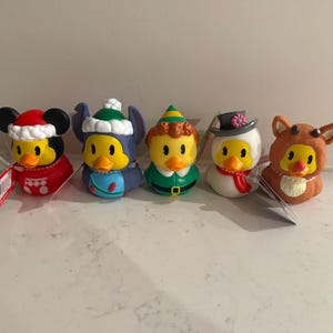 Puede incluir: Un conjunto de cinco patos de goma vestidos con disfraces navideños. El primero lleva un gorro y traje de Papá Noel rojo, el segundo un gorro de invierno azul, el tercero un traje de elfo verde, el cuarto un sombrero de copa gris y el quinto un reno.