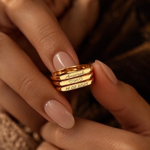 Könnte beinhalten: Ein Stapel aus drei goldfarbenen Ringen mit flachen, rechteckigen Oberflächen. Der oberste Ring ist mit dem Namen Ameerah graviert, der mittlere Ring zeigt zwei Herzen und ein Unendlichkeitssymbol, der untere Ring trägt das Datum 14.02.2025.