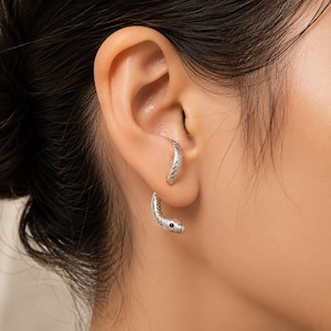 May include: A silver-toned ear cuff shaped like a snake, featuring a textured scale pattern along its body and a small, dark, circular accent representing the eye. The piece curves around the cartilage and lobe of the ear in a continuous design.