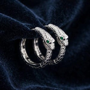 May include: A pair of silver hoop earrings shaped like snakes with textured scales and small green gemstone eyes. The snake heads meet the hoop body, creating a continuous circular design with a polished metallic finish and detailed engravings.