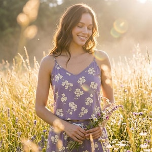 May include: A lavender-coloured sleeveless top featuring a repeating pattern of small white flowers with thin shoulder straps and a V-neckline, worn as part of a matching two-piece set in a sunlit field of wildflowers and tall grasses.