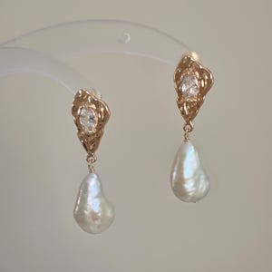 May include: A pair of gold-toned drop earrings featuring a textured, irregular metal top piece set with a clear marquise-cut stone. A single white baroque pearl hangs from each metal component, suspended by a small gold-toned jump ring.