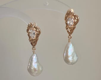 May include: A pair of gold-toned drop earrings featuring a textured, irregular metal top piece set with a clear marquise-cut stone. A single white baroque pearl hangs from each metal component, suspended by a small gold-toned jump ring.