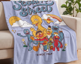 May include: A light purple throw blanket featuring the Sesame Street logo and characters including Big Bird, Elmo, Cookie Monster, Bert, Ernie, and Grover. The design includes a yellow sun, a rainbow, flowers, and the text Since 1969.
