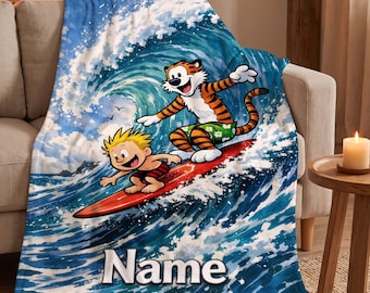 May include: A soft fleece blanket featuring a colourful illustration of a cartoon boy and a tiger surfing on a red surfboard amidst large blue ocean waves. The word Name is printed in bold white letters at the bottom of the blanket design.