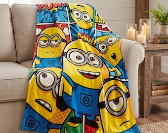 May include: A soft throw blanket featuring a colourful comic book style print of yellow Minion characters in blue overalls. The design includes various expressions and action text bubbles like POW and BAM, set against a vibrant grid pattern.