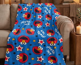 May include: A blue fleece throw blanket featuring a repeating pattern of Elmo faces, white and red stars, yellow stars, and the text "AW, YEAH!" printed in white. The blanket has a soft texture and covers the surface of a neutral-coloured sofa.