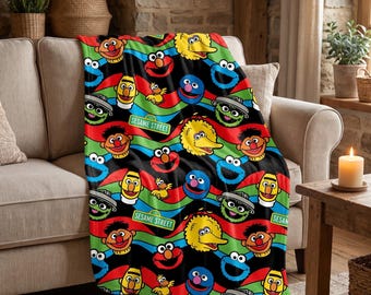 May include: A soft throw blanket featuring a repeating pattern of Sesame Street characters including Elmo, Big Bird, Cookie Monster, Bert, Ernie, and Oscar the Grouch, set against a background of wavy red, green, and blue stripes with street signs.