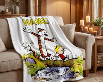 May include: A white fleece blanket featuring a colourful graphic of a cartoon tiger and a young boy walking across a log over a stream. The word NAME is printed in large yellow block letters at the top of the blanket above the cartoon characters.