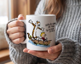 May include: A white ceramic mug featuring a colourful illustration of a tiger and a young boy walking on a log. The text printed on the mug reads, "HOLD ON TO THE GOOD STUFF." The design includes orange, black, and red accents on the characters.