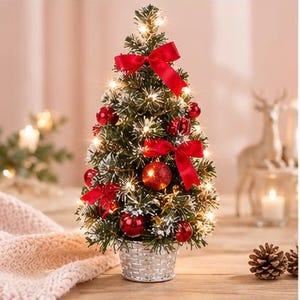 Puede incluir: Un árbol de Navidad artificial en miniatura con ramas verdes espolvoreadas de nieve, iluminado por luces blancas cálidas. El árbol está decorado con esferas rojas brillantes y tres lazos de tela roja, todo dentro de una cesta plateada.