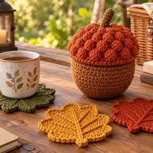 May include: A crochet acorn-shaped lidded container in orange and brown, accompanied by three leaf-shaped crochet coasters in green, yellow, and orange. A ceramic mug with leaf patterns sits on the green coaster, and a notebook with text is visible.