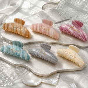 May include: Six marbled hair claw clips in various colours including tan, pink, magenta, teal, grey, and cream. Each clip features a gold-toned cursive name engraved on the surface, specifically the names Harper, Scarlett, Avery, Abigail, Edy, and Natalie.