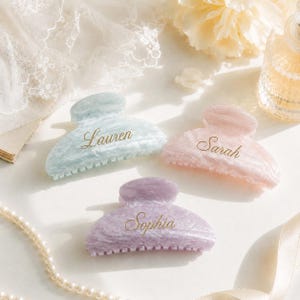 以下が含まれることがあります： ライトブルー、ソフトピンク、ラベンダーのマーブル模様のヘアクリップ3点。それぞれのクリップにはゴールドの筆記体で名前が刻印されており、ブルーには「Lauren」、ピンクには「Sarah」、ラベンダーには「Sophia」と記されています。