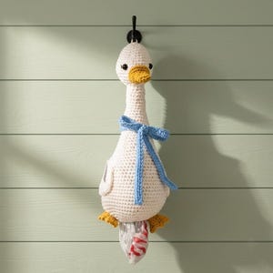 Op de afbeelding: Een crèmekleurige gehaakte gans als wandhanger met een gele snavel, kleine zwarte oogjes en een lichtblauw gehaakt lint om de nek. De gans heeft gele voeten en een opening aan de onderkant van het lichaam als dispenser voor plastic zakjes.