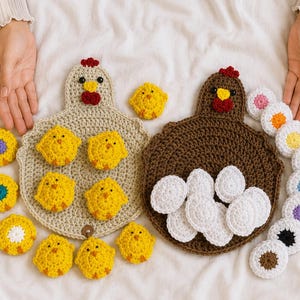May include: Two crocheted chicken-shaped mats in beige and brown, featuring red combs and yellow beaks. The beige mat is surrounded by small yellow crocheted chicks, while the brown mat holds several white crocheted egg-shaped pieces.