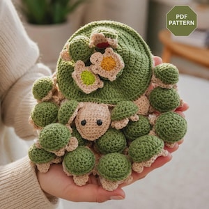 Könnte beinhalten: Eine Häkelanleitung für eine kreisförmige grüne Matte mit zahlreichen kleinen, ausgestopften Schildkrötenfiguren auf der Oberfläche. Die Schildkröten haben hellbeige Köpfe mit schwarzen Augen und grüne Panzer. Ein grünes Abzeichen mit dem Text PDF PATTERN ist sichtbar.