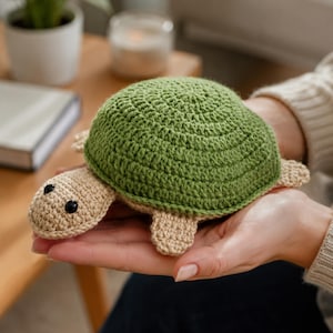 Pode incluir: Uma pequena tartaruga de crochê feita à mão com um casco verde arredondado e texturizado, além de cabeça, membros e parte inferior em bege claro. A tartaruga possui dois pequenos olhos redondos de plástico preto nas laterais.