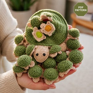 May include: A crochet pattern PDF featuring a large green turtle shell base adorned with multiple small, stuffed green and beige turtles. The central turtle has a beige head with black eyes, and the shell is decorated with small, colorful crocheted flowers in yellow, green, and white.