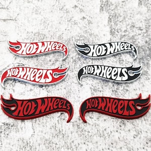 Könnte beinhalten: Sechs Hot Wheels Logo-Embleme in zwei Spalten angeordnet. Oben links rot mit weißem Text, oben rechts schwarz mit weißem Text, mittig links weiß mit rotem Text, mittig rechts weiß mit schwarzem Text und die unteren beiden sind rot mit schwarzem Text.