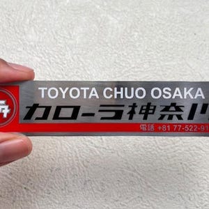 Op de afbeelding: Een rechthoekig metalen naamplaatje met een geborstelde zilveren afwerking en een rode horizontale streep. Het toont de tekst TOYOTA CHUO OSAKA, Japanse karakters, het telefoonnummer +81 77-522-9111 en een rond rood Toyota-logo.