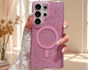 May include: A pink smartphone case featuring a glittery, speckled finish and a circular magnetic charging ring on the back. The case includes a raised rectangular camera cutout section with multiple lens openings, designed for a specific mobile device.